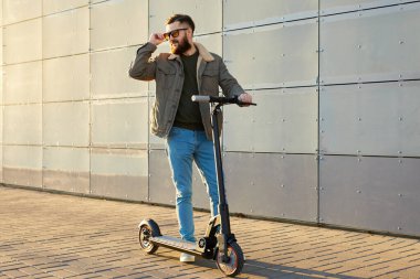 Duvarın yanında elektrikli scooterlı adam. Ekolojik ulaşım konsepti. Güneş gözlüklü şık erkek e-scooter 'da poz veriyor..