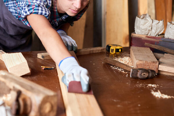 Wood grinding process by sandpaper. Female carpenter using sandpaper on a piece of timber for furniture. Woman sanding a board.