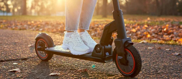 若い女性は 電動スクーターや電子スクーターで日没で街や公園を発見します スポーツスニーカーの女性の足は電動スクーターに立つ 生態系と都市交通に乗って女の子 — ストック写真