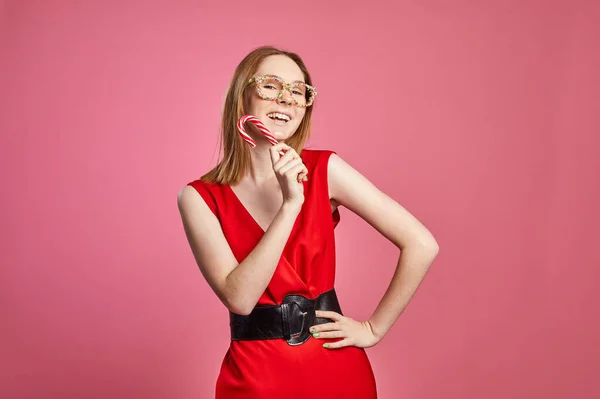 Krásná sexy mladá dívka v červených šatech a sladké sypání brýle drží slavnostní cukrovou třtinu v ruce na červeném pozadí. Stock Obrázky