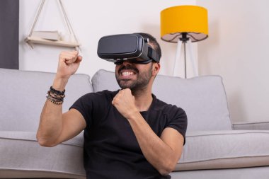 VR gözlüklü genç adam evinde koltukta heyecanlı jestler, gelecek teknolojisi, eğitim video oyunları.
