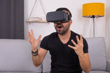 VR gözlüklü genç adam evinde koltukta heyecanlı jestler, gelecek teknolojisi, eğitim video oyunları.