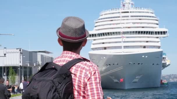 Young man tourist with hat and backpack looking at beautiful sea view with huge cruise liner behind — Stockvideo