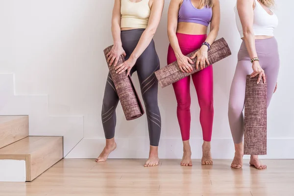 Tres mujeres jóvenes sosteniendo esterilla de yoga en ropa deportiva colorida en estudio blanco, profesor de yoga, entrenamiento —  Fotos de Stock