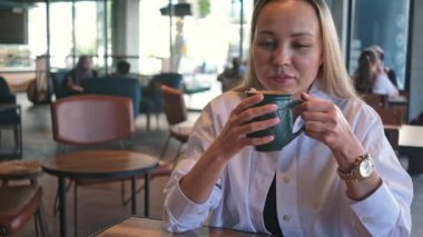 Genç bir kadın kafede oturup kahve içiyor ve dışarıda dinleniyor.