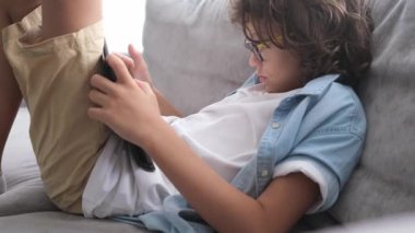 Gözlüklü, kıvırcık saçlı, kanepede oturan ve dijital tablette oyun oynayan tatlı bir çocuk. Evde dizüstü bilgisayarla oynayan çocuğun portresi. 