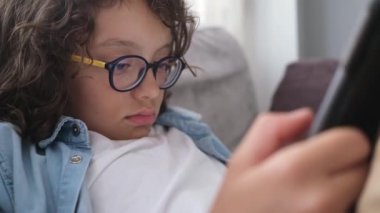 Gözlüklü, kıvırcık saçlı, kanepede oturan ve dijital tablette oyun oynayan tatlı bir çocuk. Evde dizüstü bilgisayarla oynayan çocuğun portresi. 