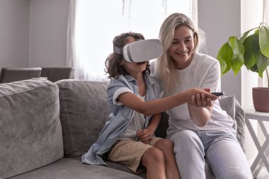 Anne, oğlunun sanal gerçeklik kulaklığı takmasına yardım ediyor. Oturma odasında sanal gerçeklik oyunları oynayarak eğleniyor.