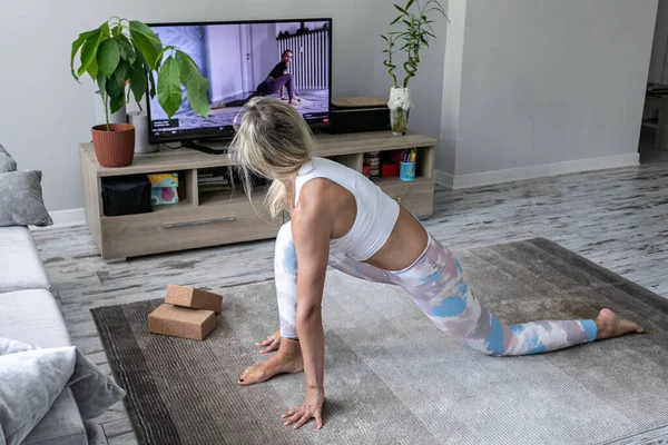 Evde yoga egzersizi yapan genç bir kadın TV ekranında öğretmenlik dersleri izliyor.