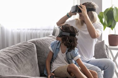 Anne ve oğlu sanal gerçeklik kulaklığı takıyor evdeki oturma odasında sanal gerçeklik oyunları oynayarak eğleniyorlar.