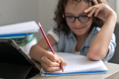 Kıvırcık saçlı, gözlüklü, uzaktan kumandalı, sevimli okul çocuğu. Dijital tablet kullanan konsantre çocuk, video dersinde öğreniyor..