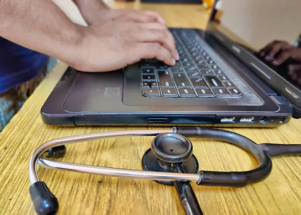Bilgisayarlı bir steteskop fotoğrafı, arka planda çekilmiş. İçerde çekilmiş. İnternette araştırma yapmak için dizüstü bilgisayar kullanan bir doktor resmedilmiş. Görüntü bulanık arkaplanlı