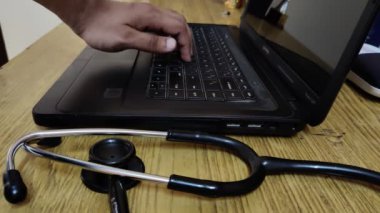Arka planda dizüstü bilgisayarı olan bir masaya yerleştirilmiş steteskop görüntüsü, klavyeyi kullanan kişi. Görüntü, doktorların teknolojinin kullanımını tasvir etmeye çalışıyor