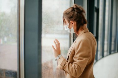 Ofiste maske takan genç bir kadın. Koronavirüs. Salgın. Covid - 19 yaşında. Pencereden bakan genç bir kadın..