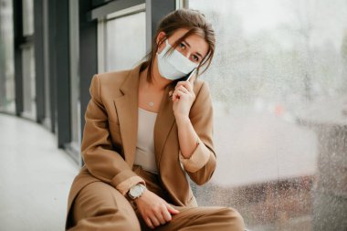 Ofiste maske takan genç bir kadın. Koronavirüs. Salgın. Covid - 19 yaşında. Pencereden bakan genç bir kadın..