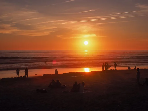 Portekiz 'de güzel bir gün batımı, Carcavelos plajı. Atlantik Okyanusu kıyısındaki karanlık insan siluetleri. İnsanlar dinleniyor, yürüyor ve sörf yapıyor..