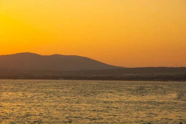 Akşam gökyüzü dağın arkasındaki batan güneşle aydınlanıyor. Denizin ve dağın üzerindeki manzara. Denizin üzerinde muhteşem bir gün batımı. Boşluğu kopyala.