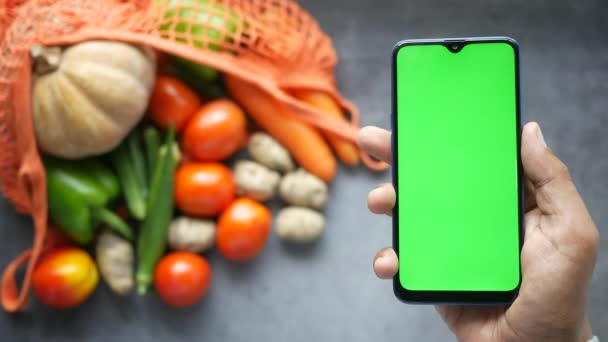 Vrchní pohled na člověka ruka drží chytrý telefon s čerstvou zeleninou na stole — Stock video