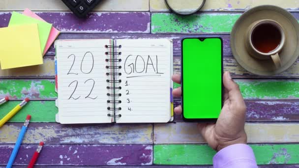 2022 novas metas ano no bloco de notas e segurando telefone inteligente na mesa — Vídeo de Stock