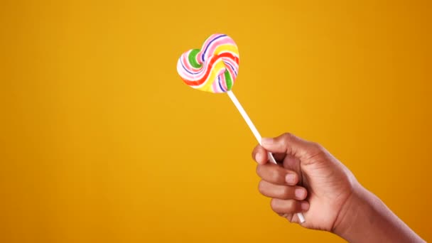 Niño mano mantenga arco iris corazón forma piruleta sobre fondo amarillo — Vídeo de stock