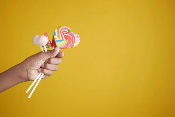 Child holding rainbow heart shape lollipop against yellow background — Stockfoto