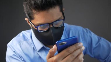 Kapalı alanda akıllı telefon kullanan maskeli genç bir adam. 