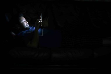 Genç adam geceleri akıllı telefon kullanarak yatakta oturuyor. 