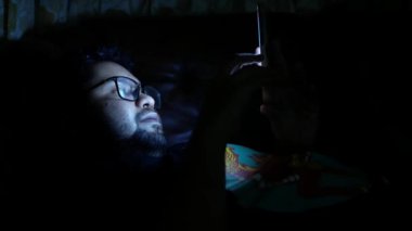 Genç adam geceleri akıllı telefon kullanarak yatakta oturuyor. 
