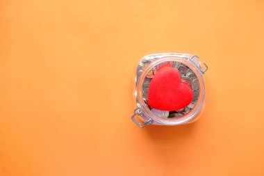 Kırmızı kalbin düz kompozisyonu ve turuncu arka plandaki kavanozdaki bozuk paralar 