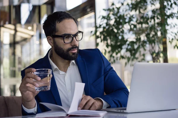 Handsome confident arabic businessman planning start up project, holding glass of water in modern office. Pensive middle eastern man watching training courses, working online sitting at workplace