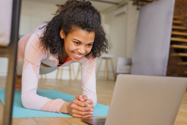 Güzel gülümseyen fitness kadını dizüstü bilgisayar izleyip, evde tahta pozlar vererek egzersiz yapıyor. Spor, sağlıklı yaşam tarzı konsepti 
