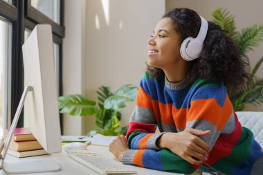 Evde çalışan bilgisayar planlama projesini kullanarak gülümseyen serbest çalışan biri. Kulaklık takmış güzel Afro-Amerikan kadın portresi iş yerinde oturup pencereye bakıyor.