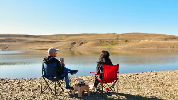年轻的高加索夫妇坐在可折叠的椅子上 一边在室外的阳光下喝咖啡 一边带着宁静的湖景背景旅行 浪漫情侣户外的概念 — 图库视频影像
