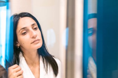Pretty woman with fresh morning face in white robe looks to the mirror suspiciously. Vertical background image