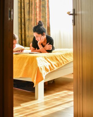 Genç anne ya da bebek bakıcısı yatağa uzanmış ve çocuğa ödevi olan okul defterini okumasında yardım etmiş. Yatak odasında oturmuş. Şefkatli anne anaokulu çocuğuna dersi açıklar. Anne-baba, oğluyla evde ders çalışıp birlikte ev ödevi yapıyor.