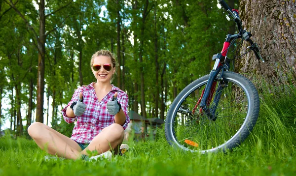 Giovane ciclista seduto nell'erba e mostrando i pollici in alto gesto — Foto Stock