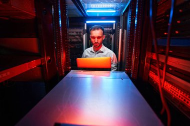 Sunucu rafının önünde dizüstü bilgisayarı olan teknisyen