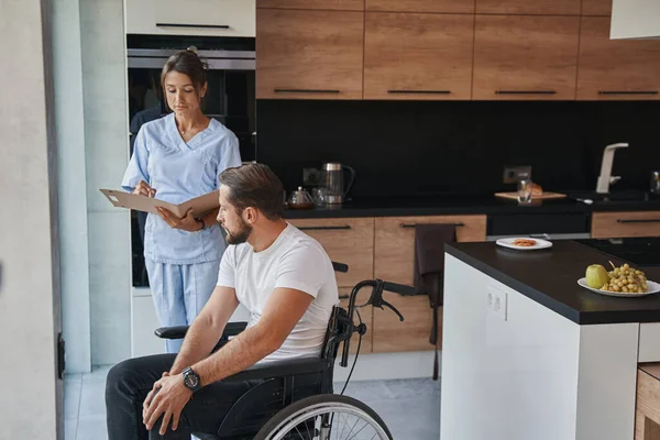 Красивый врач в форме и мужчина, который в инвалидной коляске беседует на домашней кухне — стоковое фото