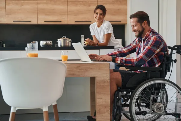 Гарний кавказький самець, який користується інвалідним візком, працюючи на ноутбуку на сучасній кухні. — стокове фото