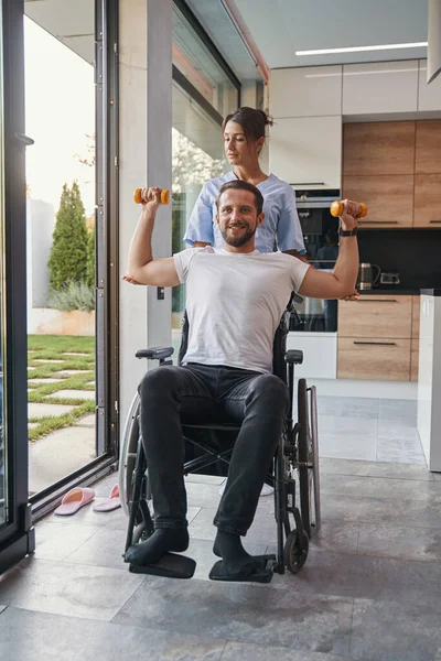 Чарівний позитивний чоловік, який використовує гантелі та інвалідне крісло на сучасній кухні — стокове фото