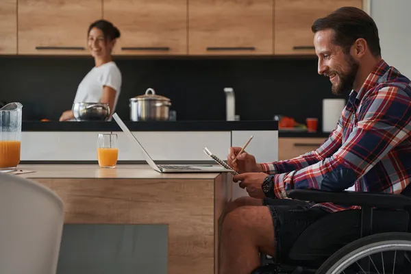 Feliz adorável deficiente masculino escrever algo no bloco de notas ao usar laptop em casa apartamentos — Fotografia de Stock