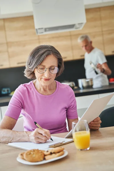 Envelhecimento mulher escrevendo no papel enquanto toma café da manhã — Fotografia de Stock