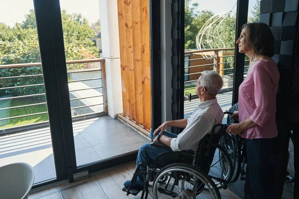휠체어를 타고 창문으로 마당을 바라보고 있는 장애인 남자 — 스톡 사진