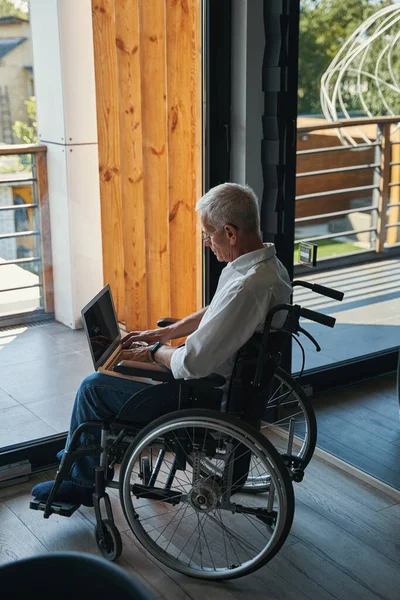 Mężczyzna na wózku inwalidzkim pracujący na laptopie w pobliżu okna domu — Zdjęcie stockowe