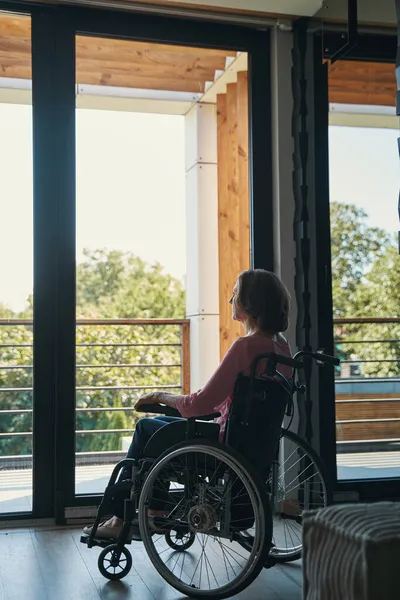 Niepełnosprawna kobieta na wózku inwalidzkim gapiąca się w okno — Zdjęcie stockowe
