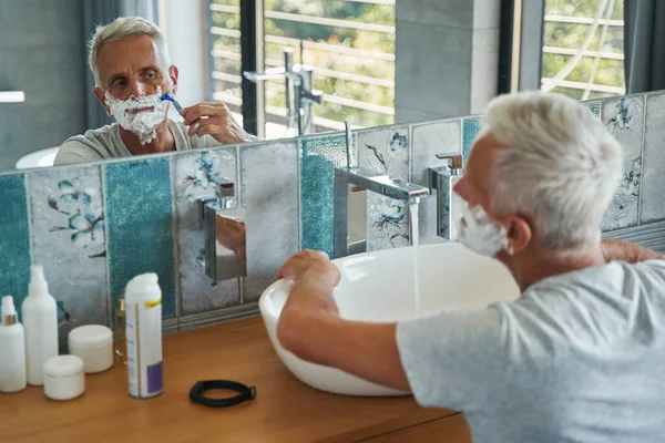 Старший мужчина удаляет крем для бритья с лица бритвой — стоковое фото