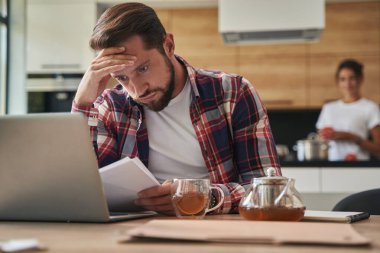 Kafası karışmış sakallı erkek, içerideki odada kağıttan bir şeyler okuyor.