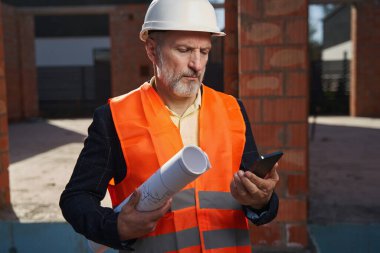 Adanmış inşaatçı inşaat planlarını getiriyor ve telefonunu kontrol ediyor.