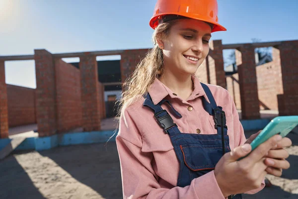 Schöne junge Frau in Bauunternehmeruniform schaut aufs Smartphone — Stockfoto