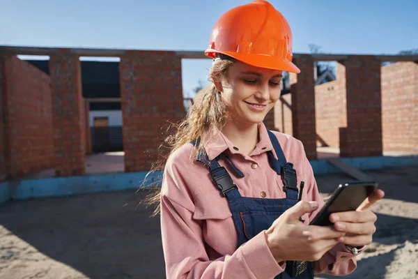 Unga kvinnliga byggare som håller smartphone i byggområdet — Stockfoto